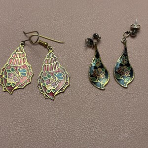 May include: Two pairs of earrings. The first pair features gold-tone metal seashell shapes with pink enamel and floral patterns. The second pair consists of gold-tone metal teardrop shapes with black enamel, featuring floral designs and butterfly accents.