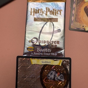 May include: A silver Harry Potter Quidditch Cup trading card booster pack containing 11 random game cards, positioned above a black Scabbers creature trading card featuring a gray rat illustration, 3 health points, and 1 damage per turn.