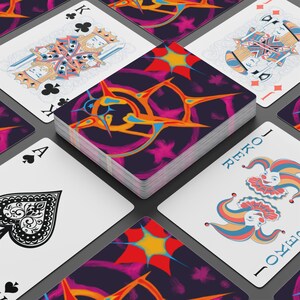 May include: A deck of playing cards with a colorful abstract design on the back. The cards are arranged with some face up, showing suits, face cards, and a joker. The deck's back features a purple background with orange, yellow, and blue shapes.