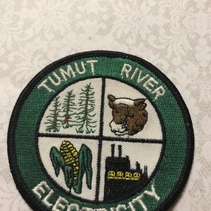 May include: Circular embroidered patch with the text "TUMUT RIVER ELECTRICITY" in white on a green and black border. The design includes images of trees, a cow, corn, and a power plant, divided into four sections.