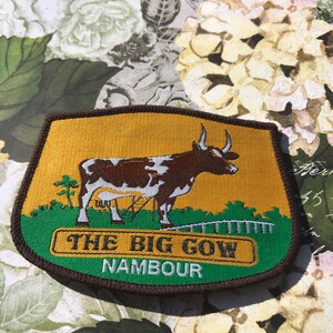 May include: A brown and white cow with horns is embroidered on a yellow patch. The patch is surrounded by a green border with the text "THE BIG COW NAMBOUR" in white.