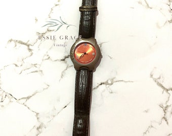 May include: A vintage wristwatch with a brown leather band and a silver-toned case. The watch face is orange with silver hands and markers. The band shows signs of wear, indicating its age. The watch is displayed on a white marble surface.
