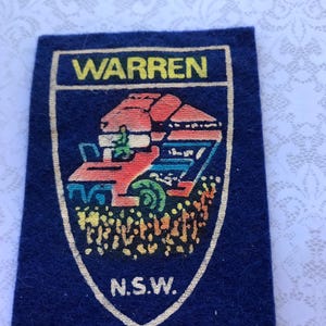 May include: A navy blue patch with a shield design. The patch features the word "WARREN" in yellow at the top, and "N.S.W." at the bottom. The shield contains a colourful illustration of a farm vehicle.