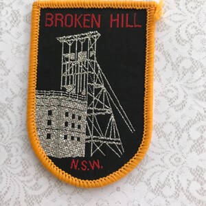 May include: A black patch with an orange border. It features the red text "BROKEN HILL" and a silver illustration of a mining structure. The text "N.S.W." is also present.