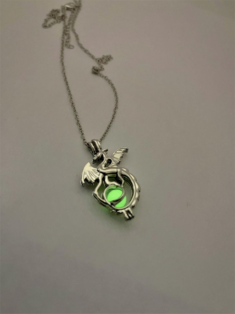 May include: A silver necklace with a dragon pendant. The dragon is holding a glowing green orb in its claws.
