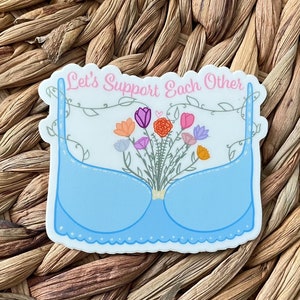 May include: A blue bra with a floral bouquet inside, surrounded by vines. The text "Let's Support Each Other" is written above the bra.