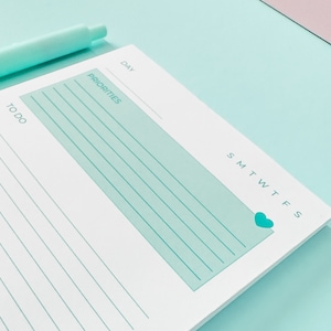 May include: A white daily planner with a light blue section for priorities and a light blue heart icon. The planner has lines for writing and a checklist with the word "to do" written next to it.