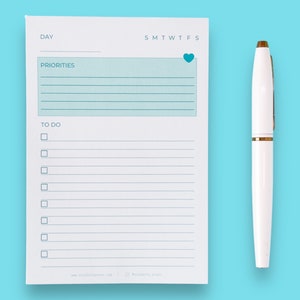 May include: A white notepad with a blue heart in the top right corner. The notepad has sections for "Day", "Priorities", and "To Do". There are lines for writing and checkboxes for the "To Do" section. A white pen with a gold accent is next to the notepad.