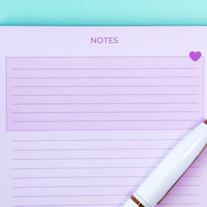 May include: A purple lined notepad with the word "NOTES" printed at the top and a small pink heart in the top right corner. A white pen with a gold accent is lying on the notepad.
