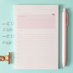 May include: A white notepad with pink lines and a pink heart in the top right corner. The notepad has the words "Day", "Priorities", and "To Do" written on it. There are also the days of the week written across the top. A pink pen and a gold binder clip are on a light blue background.