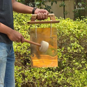 Pode incluir: Uma moldura de madeira com um gongue de cobre pendurado. Uma pessoa está segurando um martelo de madeira com uma cabeça branca e batendo no gongue.