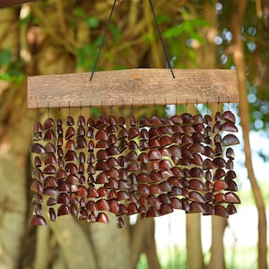 May include: A handcrafted wind chime featuring a wooden rectangular top with numerous small, brown, shell-like pieces suspended below. The shells are arranged in rows, creating a decorative and natural aesthetic. The chime hangs from a black string.