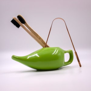 May include: A green ceramic neti pot with a wooden toothbrush and a copper tongue scraper inside.