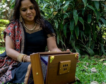 May include: A woman sits on the grass, holding a wooden harmonium with a dark blue interior. The harmonium has a small panel with six holes on the front. The woman is wearing a black blouse, a red and black shawl, and gold jewelry.
