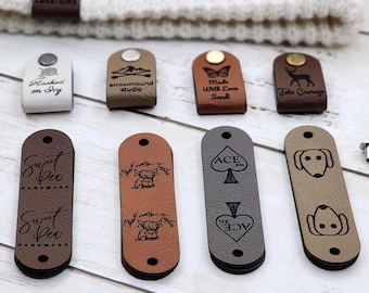 Puede incluir: Una colección de etiquetas de cuero en varios colores y diseños, incluyendo etiquetas con el texto "Sweet Pea Handmade", una escena de montaña, una mariposa y un ciervo. Las etiquetas se muestran con remaches y un gorro de punto blanco.
