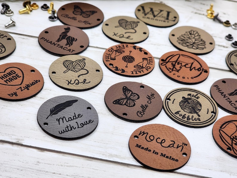 May include: Assortment of round leather labels with various designs and text. Some labels feature a butterfly, a feather, a heart, yarn, and text like "Made with Love", "Belle Moi", "Full of Jazz", "Anchor Studio", "Phantcat", "Rex", "WI Crochet", "Melo Bobbles", and "Mocean Made in Maine".