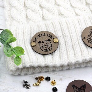 May include: A white knitted beanie with a brown leather patch that says "the little tyger" with a tiger illustration. The beanie is decorated with additional brown leather patches with a pine cone and a butterfly design.