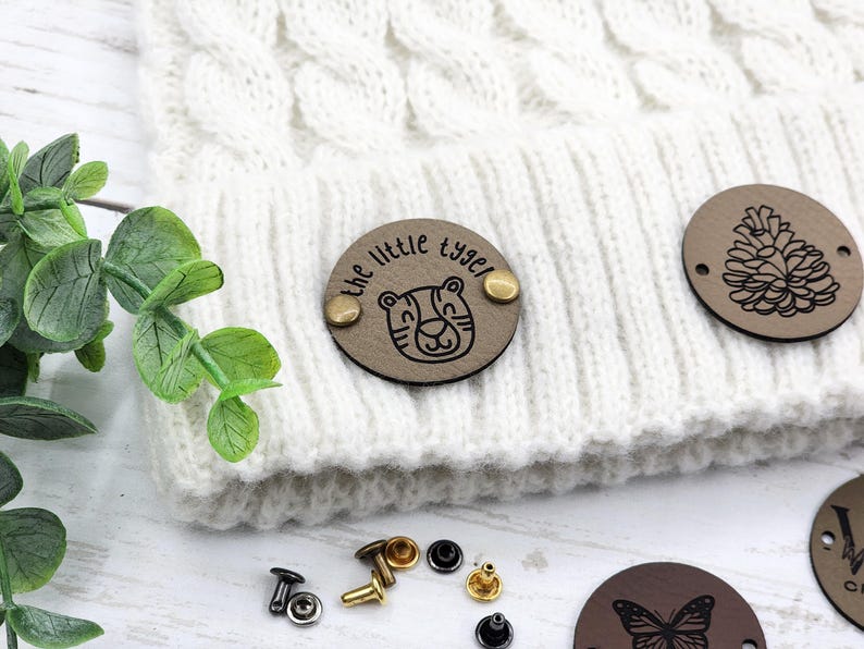 May include: A white knitted beanie with a brown leather patch that says "the little tyger" with a tiger illustration. The beanie is decorated with additional brown leather patches with a pine cone and a butterfly design.