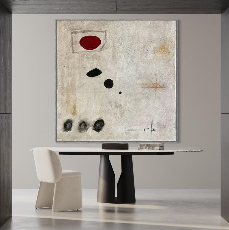 May include: Abstract painting with a red circle, black dots, and lines on a white background. The painting is framed and hanging on a wall above a table with a white top and black legs.
