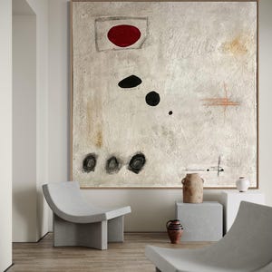 May include: Abstract painting with a red circle, black circles, and white and brown brushstrokes on a white background. The painting is hanging on a wall in a room with two concrete chairs and a concrete table.