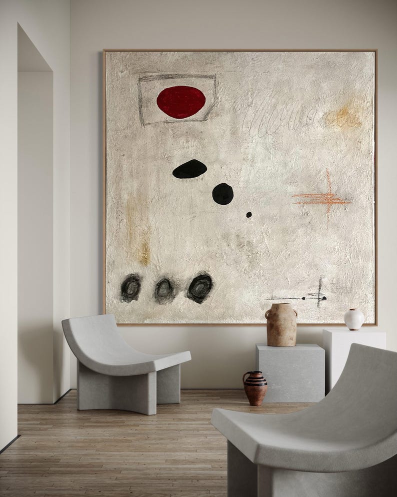 May include: Abstract painting with a red circle, black circles, and white and brown brushstrokes on a white background. The painting is hanging on a wall in a room with two concrete chairs and a concrete table.