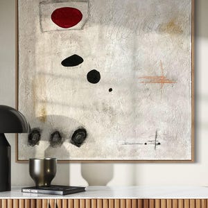May include: Abstract painting with a white background, featuring a red circle, black circles, and a brown cross shape. The painting is framed in a light wood frame.