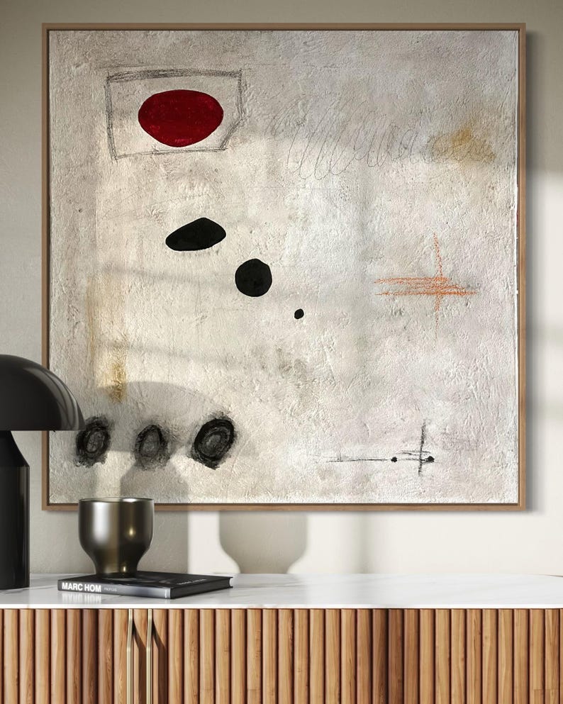May include: Abstract painting with a white background, featuring a red circle, black circles, and a brown cross shape. The painting is framed in a light wood frame.