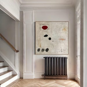 May include: A white abstract painting with a red circle, black dots, and a few lines. The painting is hanging on a white wall in a hallway with a wooden staircase and a black radiator.