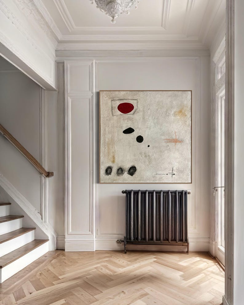 May include: A white abstract painting with a red circle, black dots, and a few lines. The painting is hanging on a white wall in a hallway with a wooden staircase and a black radiator.