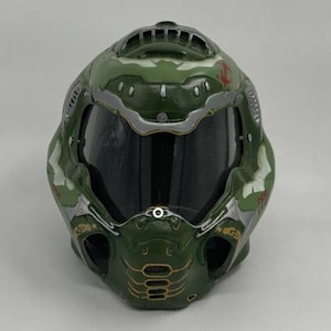 Puede incluir: Casco verde y plateado con una visera de malla negra y detalles dorados. El casco tiene un diseño de calavera con un patrón rojo y blanco en la parte superior.