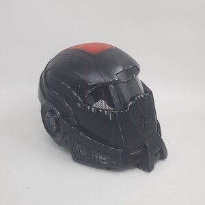 May include: Black helmet with a red stripe on top and a black visor. The helmet has a textured surface and a detailed design.