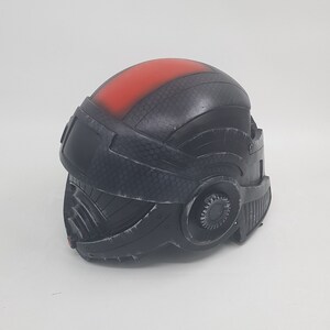May include: A black helmet with a red stripe on the top. The helmet has a textured surface and a gear-like detail on the side.