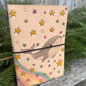 May include: A tan leather journal with a whimsical design. It features a hand-drawn whale, colorful stars, and a rainbow. The journal is secured with a black elastic band and string closure. The cover is decorated with yellow, purple, and pink stars.