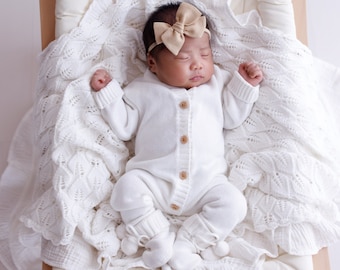 May include: A newborn baby wearing a white knitted onesie and a beige bow headband is sleeping on a white quilted pad with a white crocheted blanket.