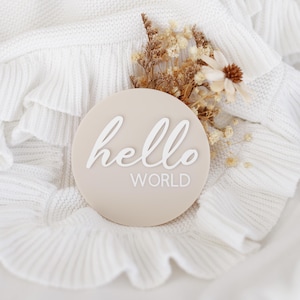 May include: A beige round sign with the text "hello world" in white lettering. The sign is on a white knitted blanket with a ruffle trim.