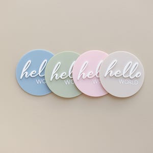 May include: Four round silicone baby announcement plaques in blue, green, pink, and beige with the text "hello world" in white.