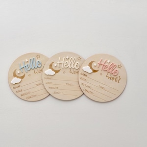 May include: Three wooden baby announcement plaques featuring a moon and cloud design. The plaques are painted in light blue, light pink, and natural wood. Each plaque has the text "Hello World" and spaces to write the baby's name, date, time, weight, and length.