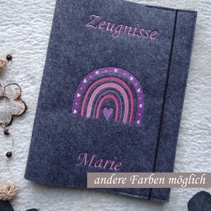 Könnte beinhalten: Eine dunkelgraue Filzmappe mit einer gestickten lila und rosafarbenen Regenbogenapplikation mit kleinen weißen Herzen. Das Wort Zeugnisse ist in Rosa darüber gestickt, und der Name Marie ist in Rosa darunter eingestickt.