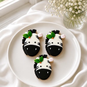 May include: Three sugar cookies shaped like zebra heads, decorated with white and black icing. Each cookie features black stripes, a black snout, small round eyes, a pink ear, and a green leaf accent with a tiny orange dot on the forehead.