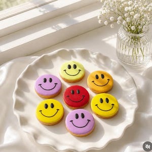 Könnte beinhalten: Sieben runde Kekse auf einem weißen Teller mit gewelltem Rand. Jeder Keks hat eine glatte, farbige Glasur in Gelb, Rot oder Lila und ist mit einem schwarzen, aufgespritzten Smiley-Gesicht verziert, das aus zwei ovalen Augen und einem Mund besteht.