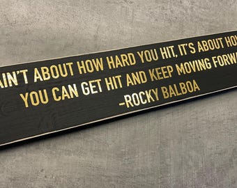 Puede incluir: Un letrero de madera negro con letras doradas que dice "IT AIN'T ABOUT HOW HARD YOU HIT, IT'S ABOUT HOW HARD YOU CAN GET HIT AND KEEP MOVING FORWARD - ROCKY BALBOA"