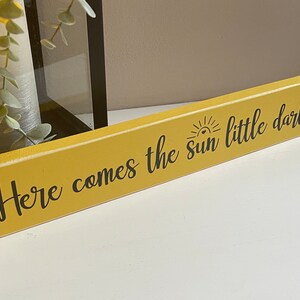 May include: A yellow wooden sign with black text that reads "Here comes the sun little darling" with a small sun graphic above the word "sun".