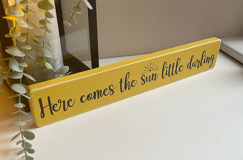 May include: A yellow wooden sign with black text that reads "Here comes the sun little darling" with a small sun graphic above the word "sun".