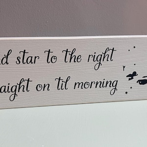Peut inclure: Un panneau rectangulaire en bois blanc avec le texte noir "Second star to the right and straight on til morning". À droite du texte se trouve une silhouette noire représentant une tour d'horloge, des étoiles et trois personnages volants.