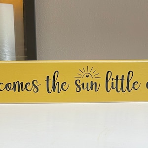 May include: A yellow wooden sign with black lettering that reads "Here comes the sun little darling". The sign has a small sun graphic with a heart in the center.