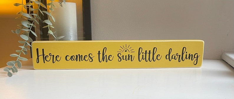 May include: A yellow wooden sign with black lettering that reads "Here comes the sun little darling". The sign has a small sun graphic with a heart in the center.