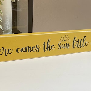 May include: A yellow wooden sign with black text that reads "Here comes the sun little darling". The sun is depicted with a heart in the center.