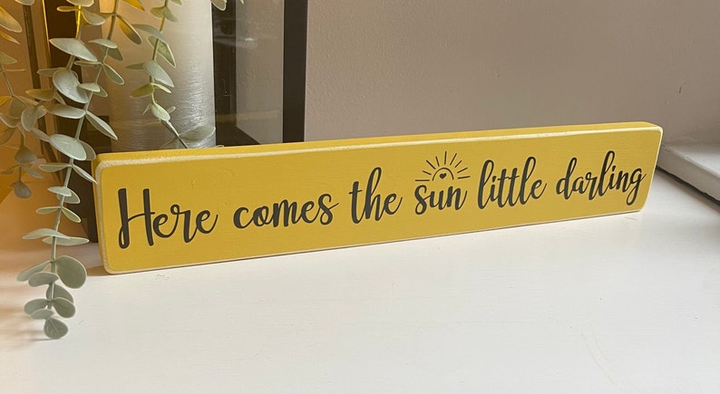 May include: A yellow wooden sign with black text that reads "Here comes the sun little darling". The sun is depicted with a heart in the center.