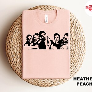 May include: Heather peach-colored t-shirt with a black graphic of six people. The shirt is folded on a woven, circular surface. The text "HEATHER PEACH" is at the bottom right. The shirt is made of a soft material.