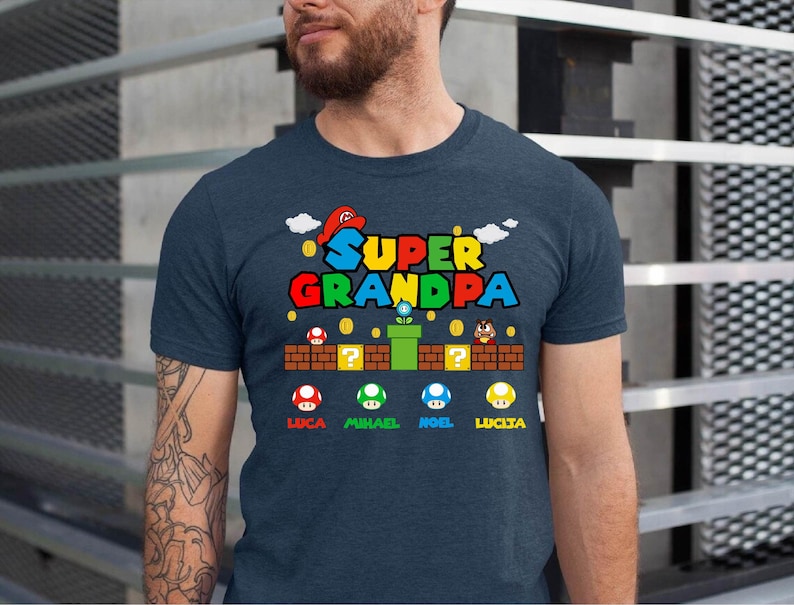 Op de afbeelding: Een donkerblauw T-shirt met de tekst "SUPER GRANDPA" in kleurrijke letters. Het ontwerp bevat een cartoonfiguur uit een videogame, een groene pijp en een bakstenen muur met vraagtekens. Er zijn vijf paddenstoelen met namen eronder: Luca, Michael, Noel, Lucia.